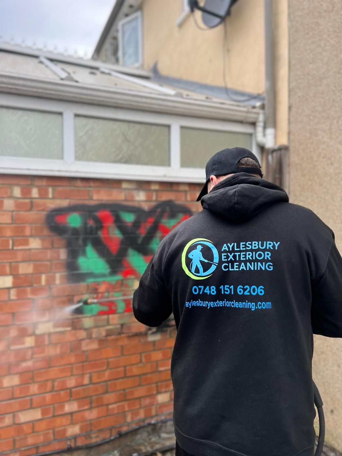 Worker using a pressure washer to remove red and green graffiti from a brick wall